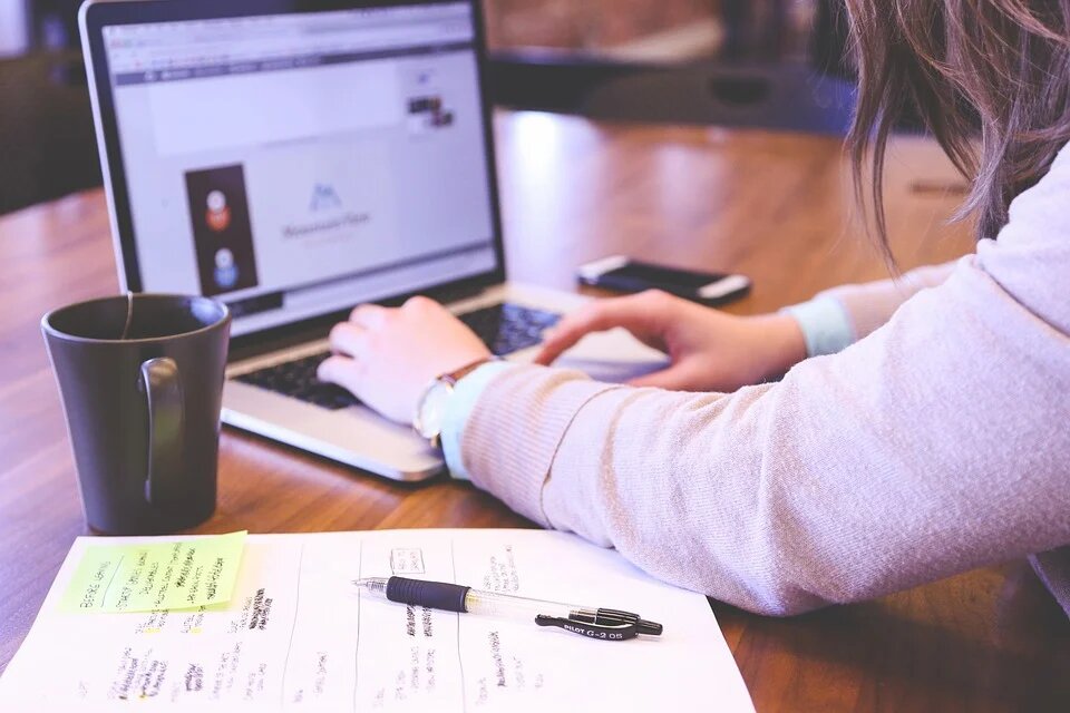 Девушка работает за ноутбуком, рядом с которым стоит чашка и лежат листки с пометками